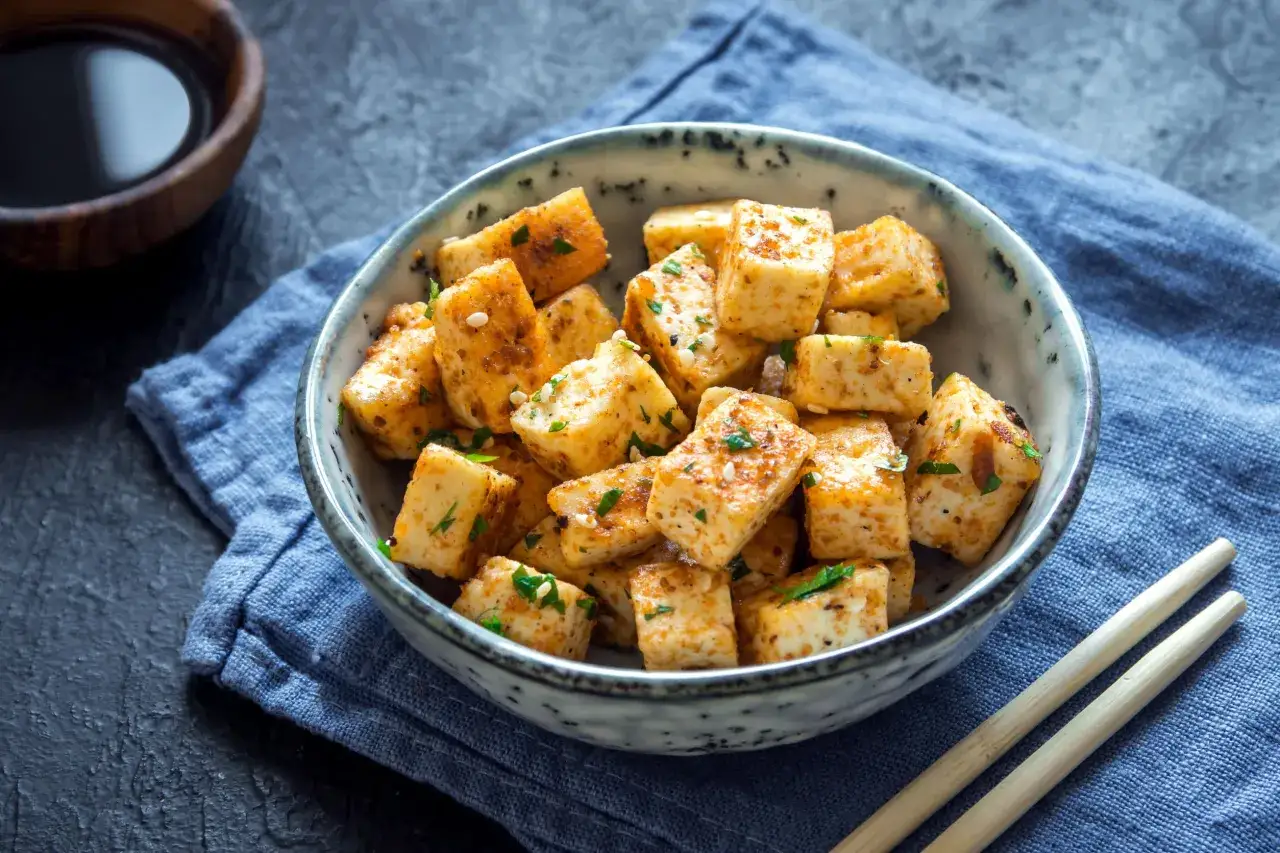 Kawałki smażonego tofu z ziołami w misce, obok pałeczki i sos.