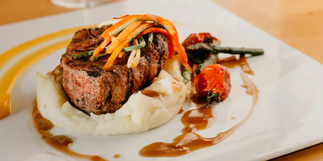 Un plat de viande grillée, purée de pommes de terre et légumes, une des spécialités d'Aurillac, servi sur une assiette blanche avec une sauce brune.