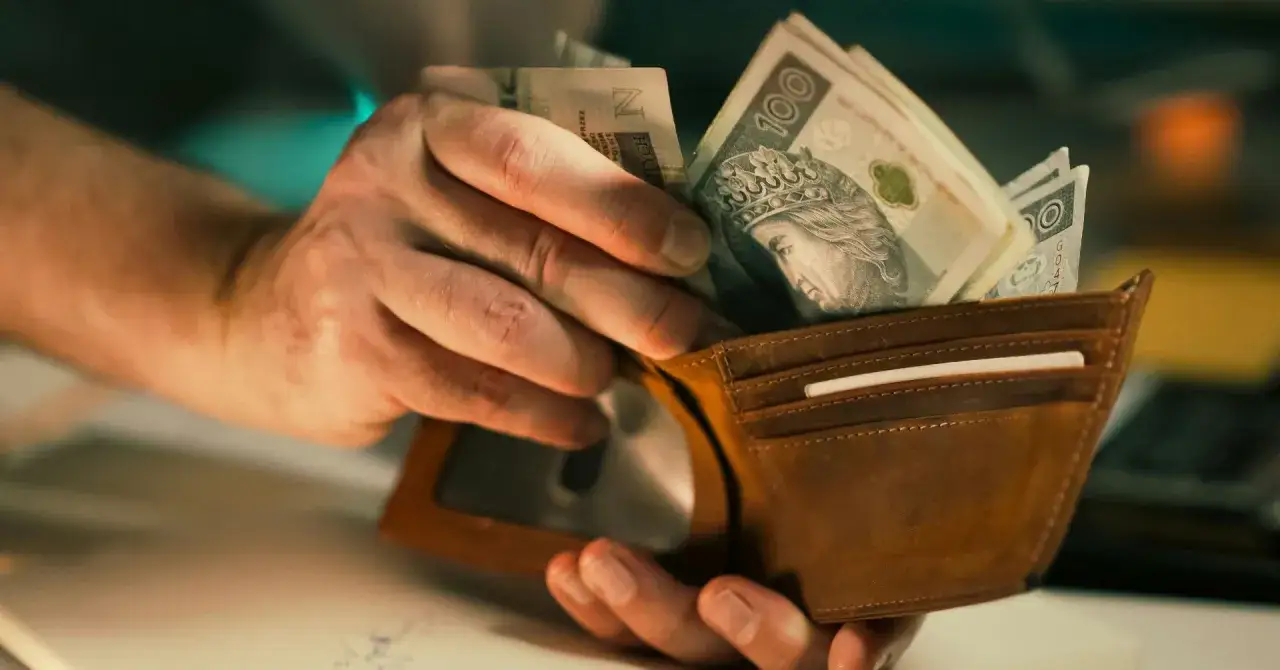 Dłonie wkładają polskie banknoty do brązowego portfela. Czy to wystarczy na minimalną stawkę godzinową netto?