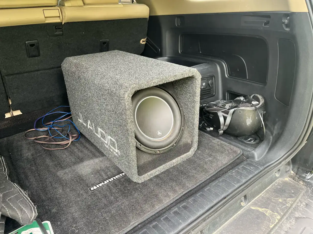 Szary subwoofer JL Audio w bagażniku samochodu, gotowy do montażu. Widać kable i akcesoria.