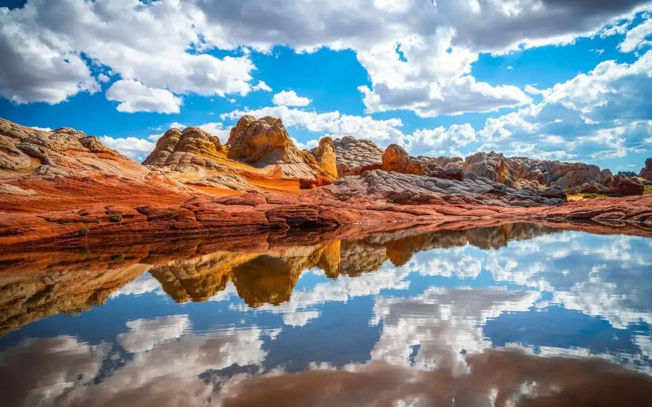 Piękne, pofałdowane skały w odcieniach czerwieni i beżu odbijają się w tafli wody wielkiej i czystej. Niebo z chmurami dopełnia krajobraz.