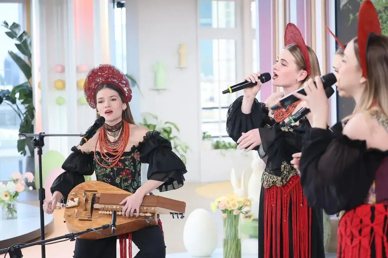 Ludowy grajek z Ukrainy gra na lirze korbowej, a obok niej dwie wokalistki śpiewają do mikrofonów.
