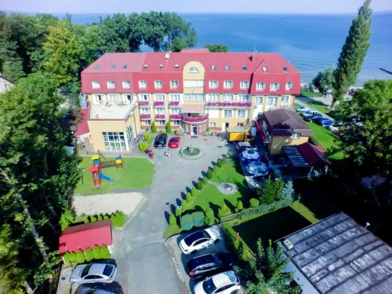 Ośrodek wypoczynkowy Jantar z czerwonym dachem, otoczony zielenią, z widokiem na morze. Na terenie plac zabaw i parking.