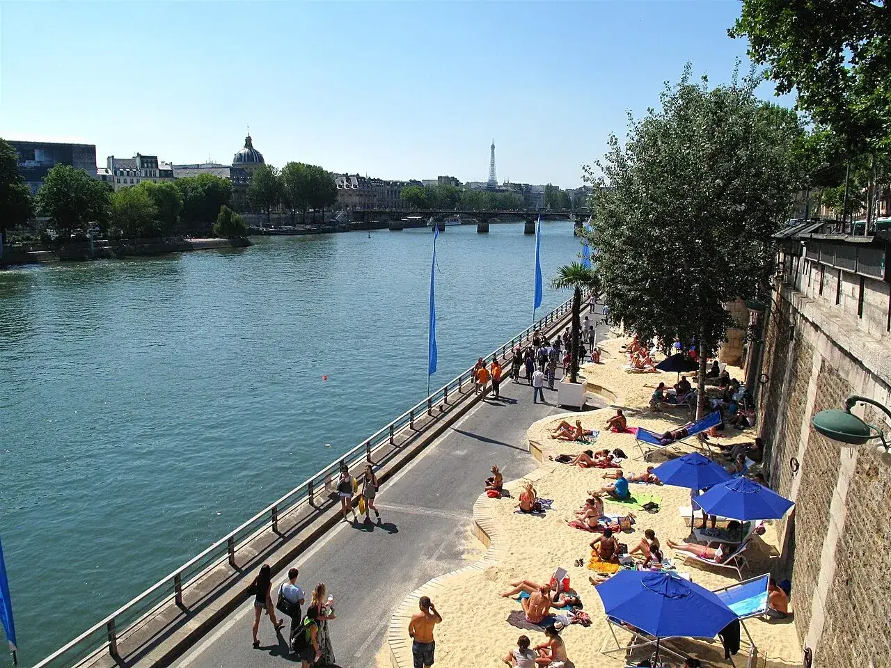 Plaża nad Sekwaną w Paryżu to świetny pomysł, co robić w Paryżu w sierpniu. Ludzie odpoczywają na piasku, pod parasolami, z widokiem na Wieżę Eiffla.