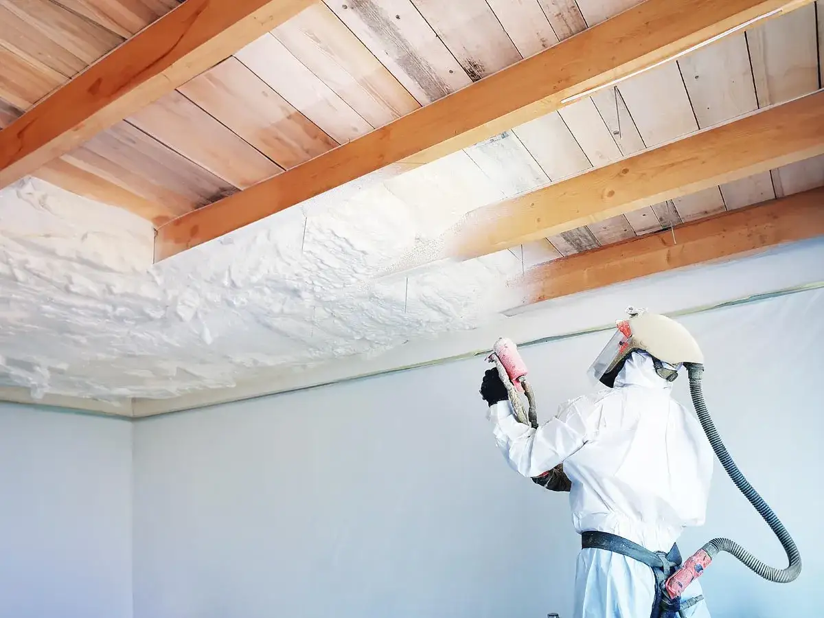 Pracownik w kombinezonie natryskuje piankę do ocieplenia poddasza na drewniane belki i deski, tworząc izolację.