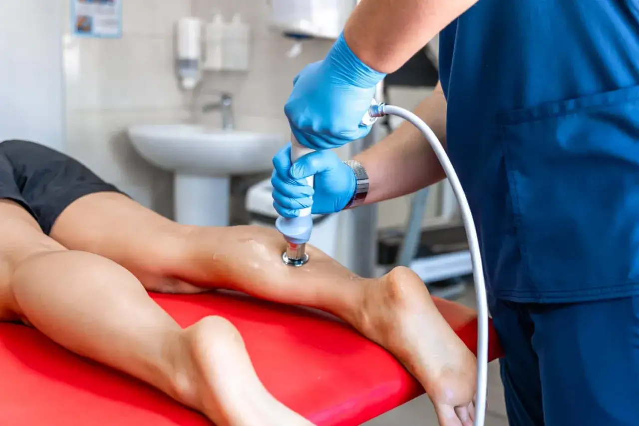 Fizjoterapeuta w niebieskich rękawiczkach wykonuje rehabilitację laserem na łydce pacjentki, co pokazuje, na czym polega ten zabieg.