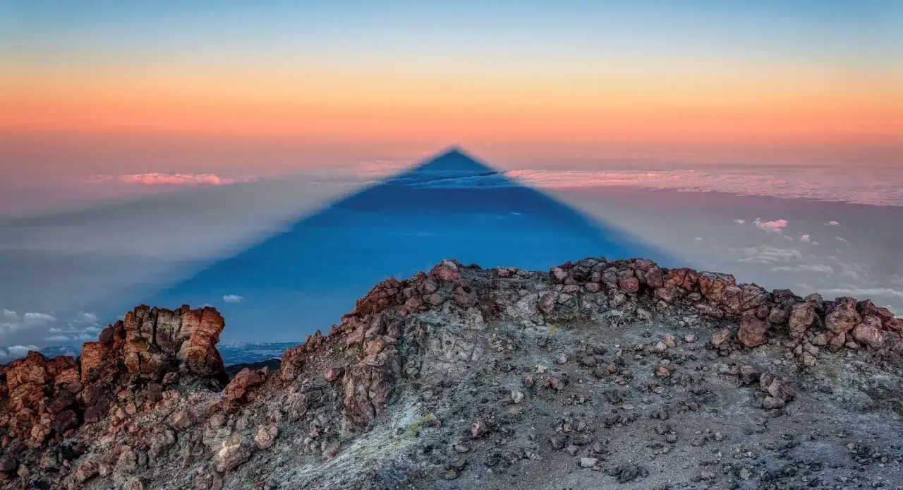Widok z góry na wulkaniczny krajobraz, gdzie cień góry tworzy idealny trójkąt na tle chmur i nieba o zachodzie słońca. Wejście na Teide to niezapomniane przeżycie.