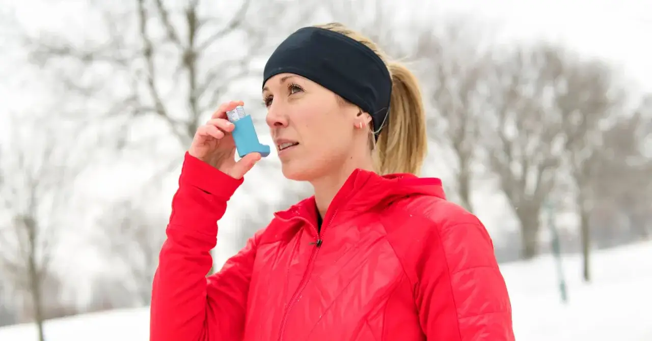 Kobieta w czerwonej kurtce używa inhalatora, by złagodzić objawy astmy wysiłkowej podczas zimowego treningu.