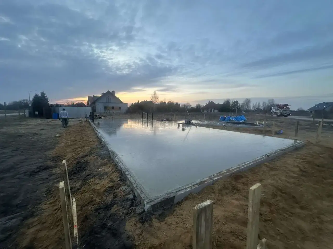 Świeżo wylana płyta fundamentowa, widać jak ile schnie płyta fundamentowa. W tle domy i zachodzące słońce.