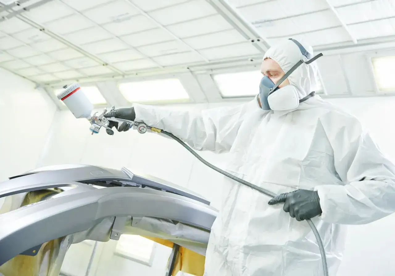 Pracownik w kombinezonie ochronnym wykonuje malowanie plastik&oacute;w w aucie, używając pistoletu lakierniczego.