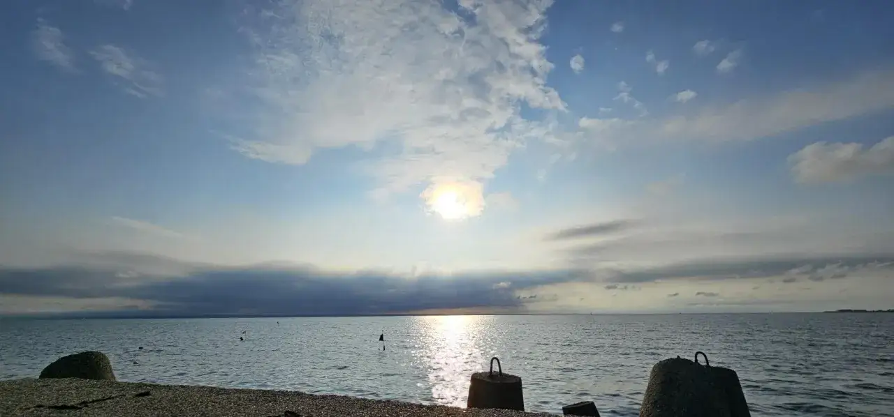 Słońce przebija się przez chmury, tworząc refleksy na spokojnym morzu. Widoczny aktualny stan morza, z betonowymi falochronami na pierwszym planie.