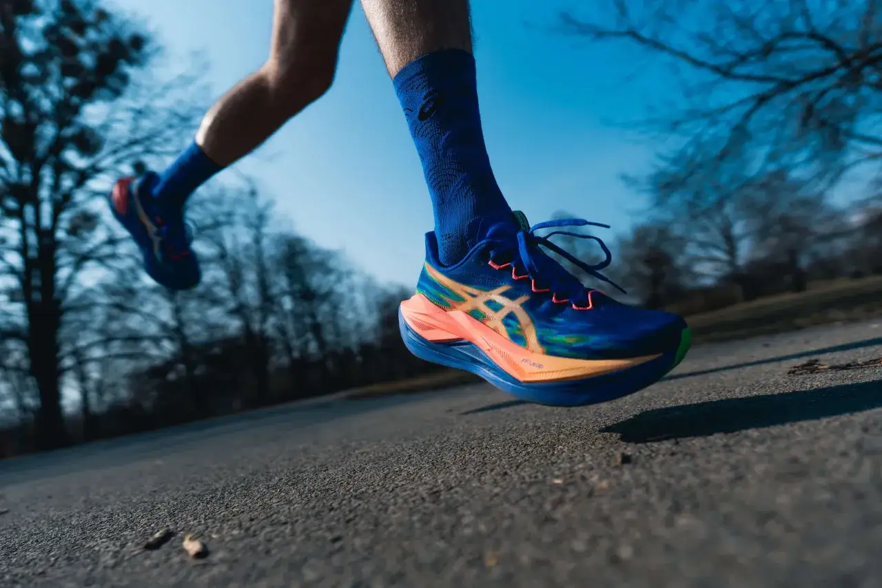 Biegacz w niebieskich skarpetkach i butach do biegania Asics, z dynamicznym designem, pokonuje asfaltową trasę wśr&oacute;d drzew.