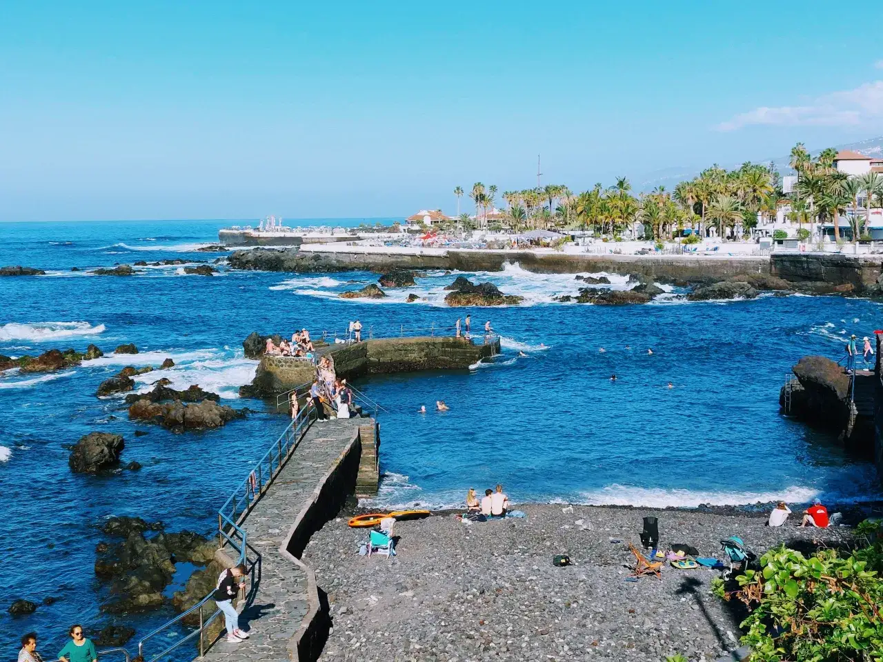 Ludzie kąpią się i opalają nad turkusowym morzem, a palmy i białe budynki tworzą tło. Czy w Hiszpanii jest morze? Tak, i to jakie!