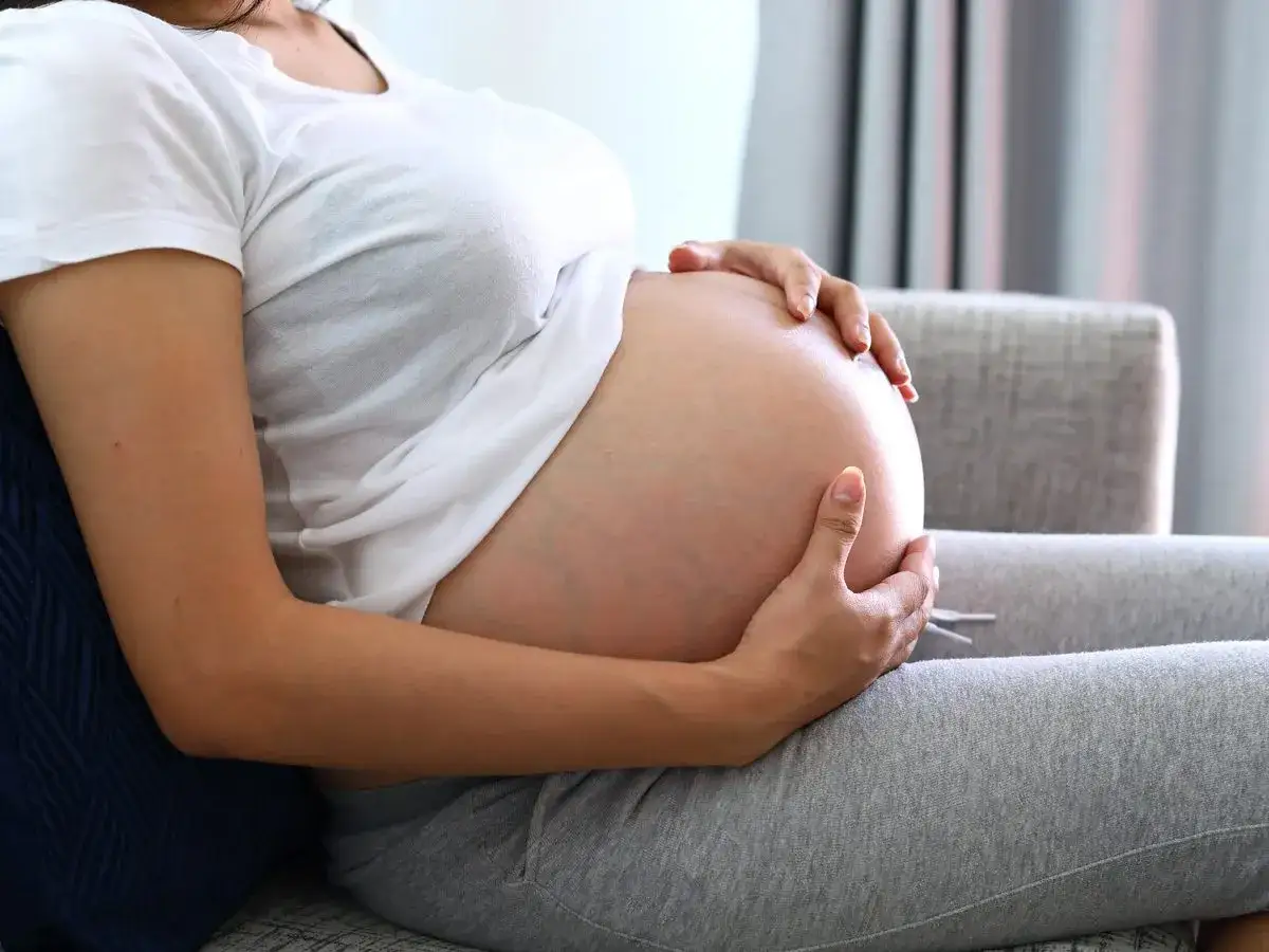 Kobieta w białej koszulce i szarych legginsach tuli duży brzuch ciążowy. Czeka na poród po terminie.