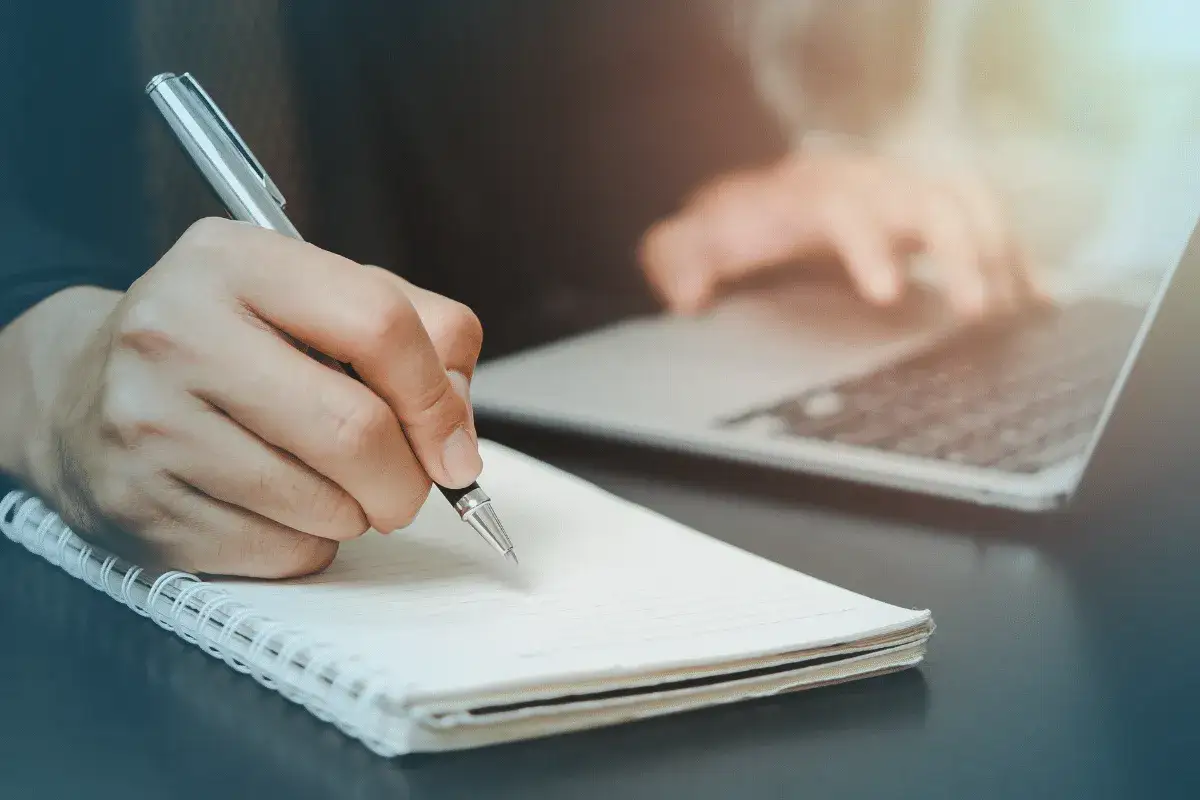 Dłoń pisze pi&oacute;rem w notatniku, obok laptop. To początek tworzenia książki, gdzie pomysły nabierają kształtu.