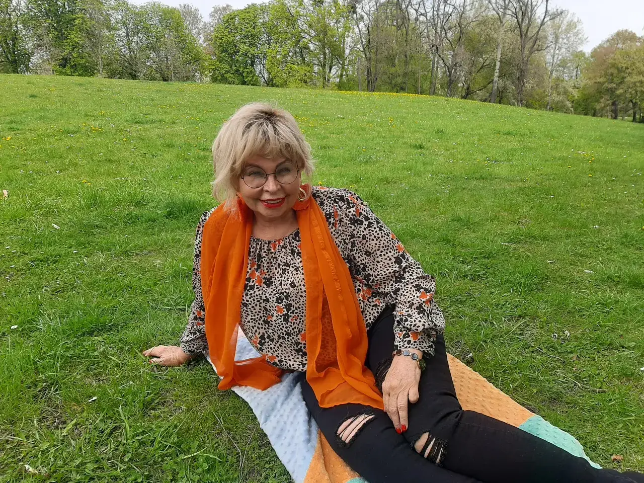 Uśmiechnięta, atrakcyjna 60 latka w pomarańczowym szalu i wzorzystej bluzce siedzi na trawie w parku.