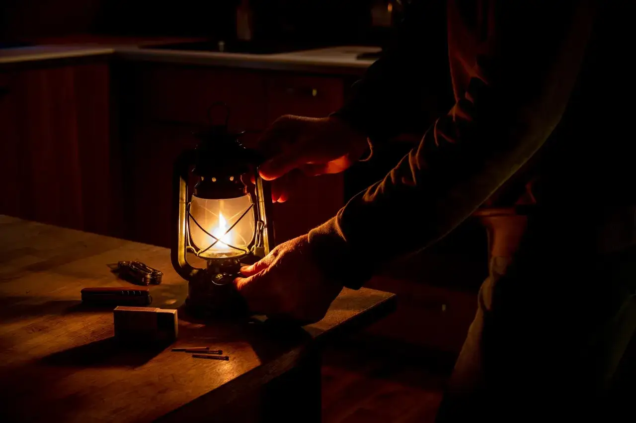 Ręce regulują płomień naftowej lampy, zapewniając oświetlenie bez prądu. Obok leżą zapałki i scyzoryk.