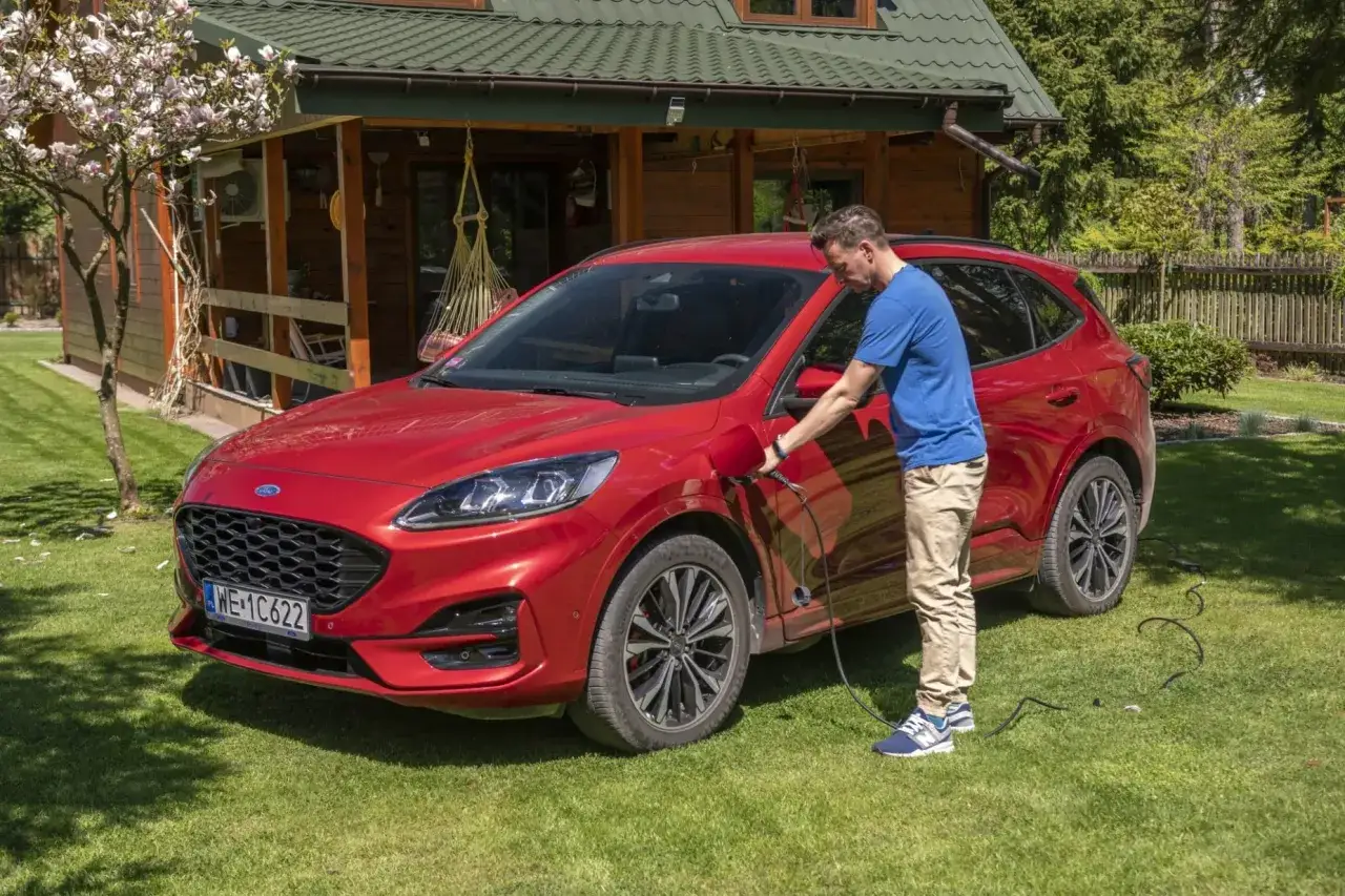 Mężczyzna ładuje czerwonego Forda Kuga. To właśnie plug-in hybrid &ndash; auto, kt&oacute;re możesz ładować z gniazdka, oszczędzając na paliwie.