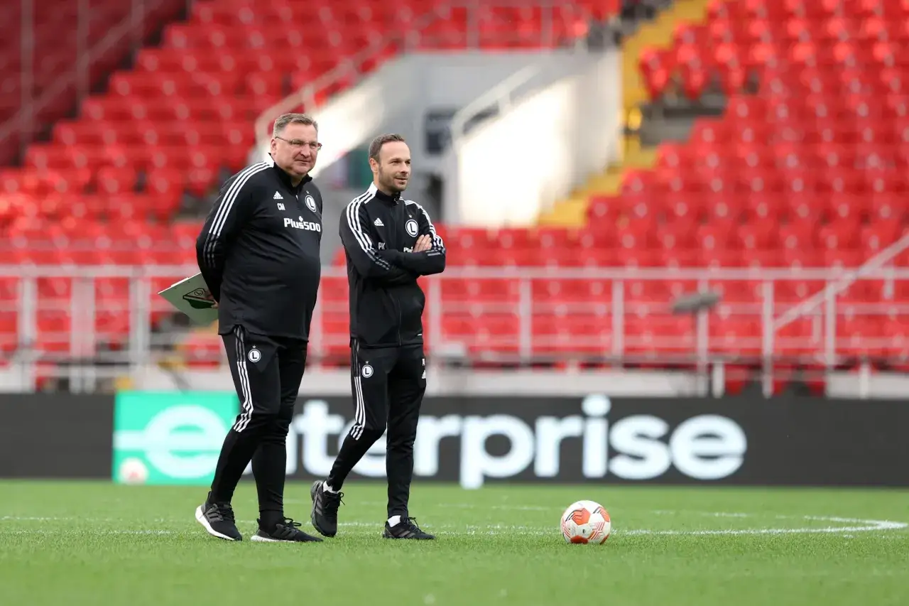 Dwóch trenerów Legii Warszawa w czarnych dresach stoi na murawie, obserwując boisko.