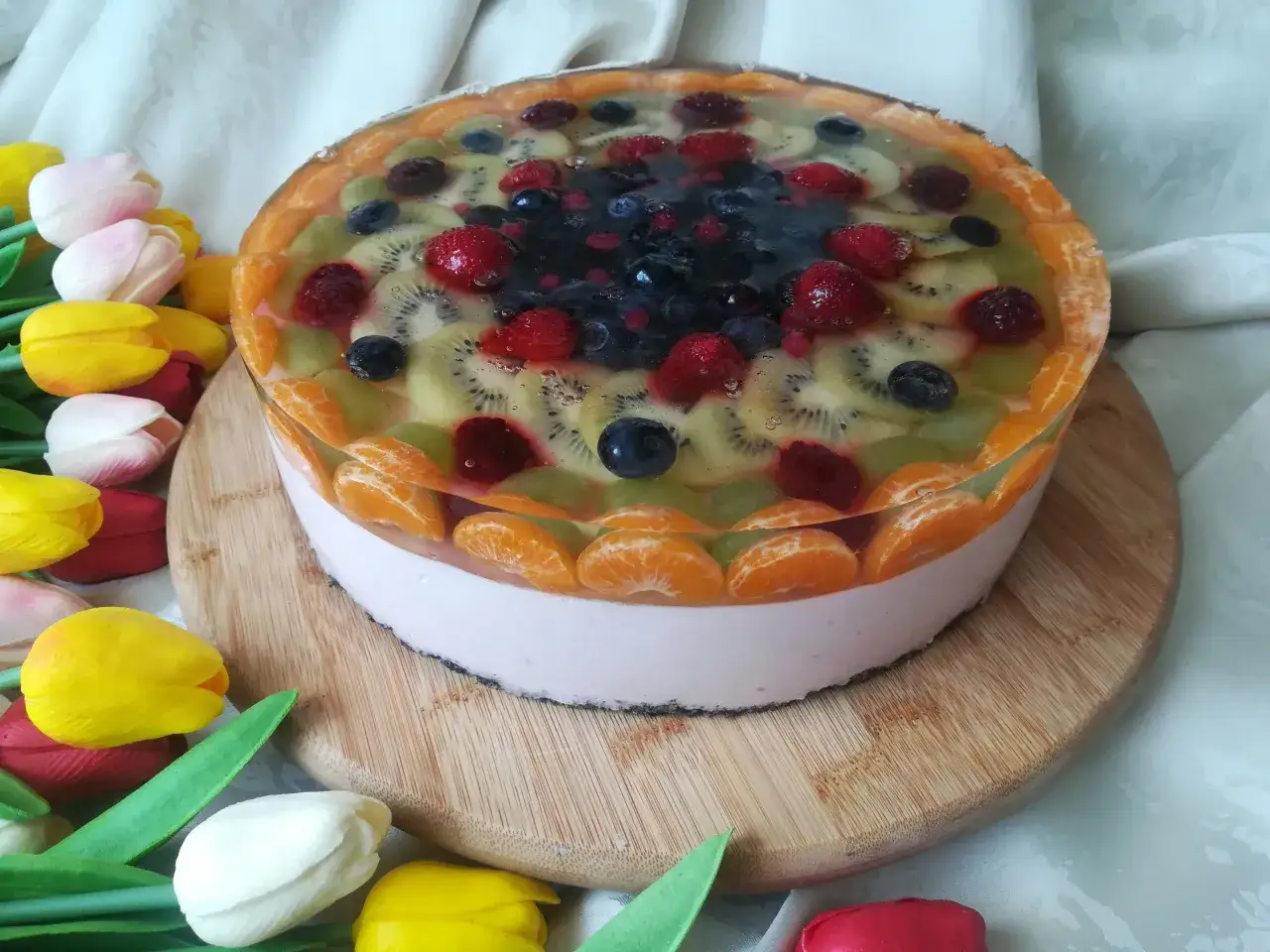 Kolorowy sernik na zimno z galaretką, ozdobiony mandarynkami, kiwi, bor&oacute;wkami i malinami.