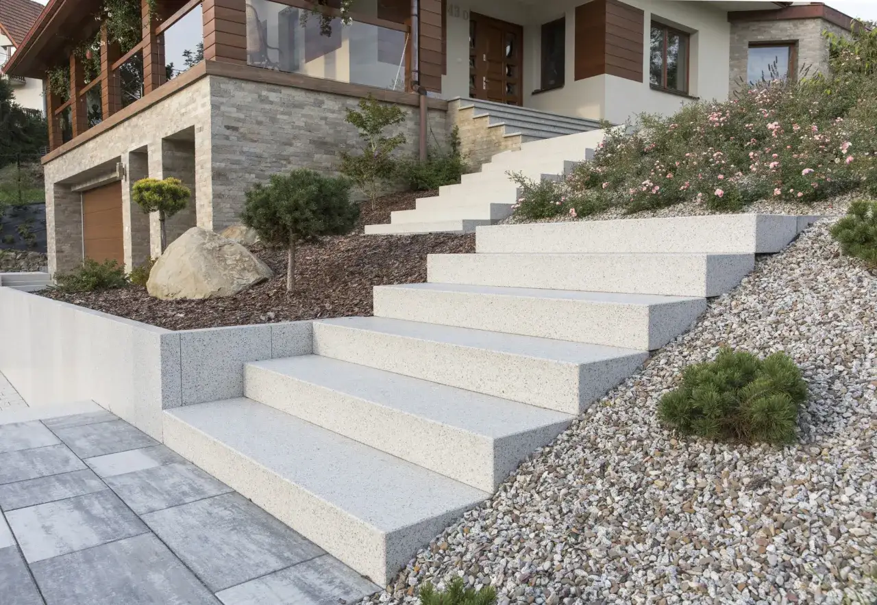 Nowe schody z bloczka betonowego prowadzą do nowoczesnego domu. Krzewy i kamyki tworzą malowniczy krajobraz.