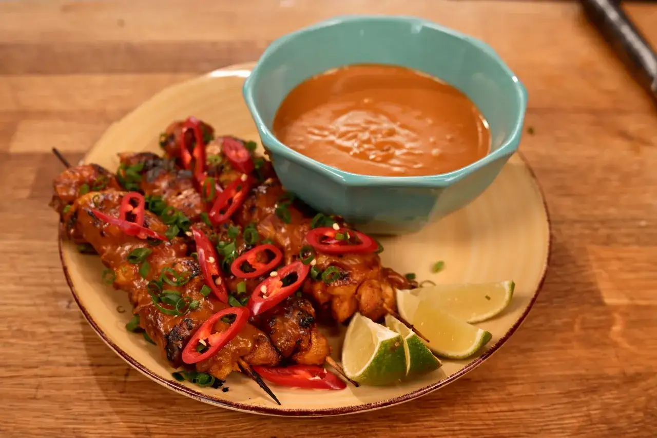 Pyszne satay z kurczaka na patyczkach, polane sosem orzechowym, ozdobione plasterkami chili i szczypiorkiem. Podane z ćwiartkami limonki.
