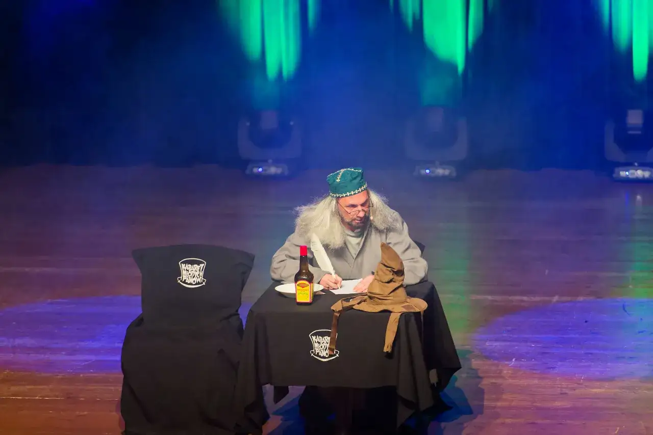 Aktor w peruce i czapce pisze pi&oacute;rem, siedząc przy stole z Tiara Przydziału. Na stole logo Kabaretu Młodych Pan&oacute;w.