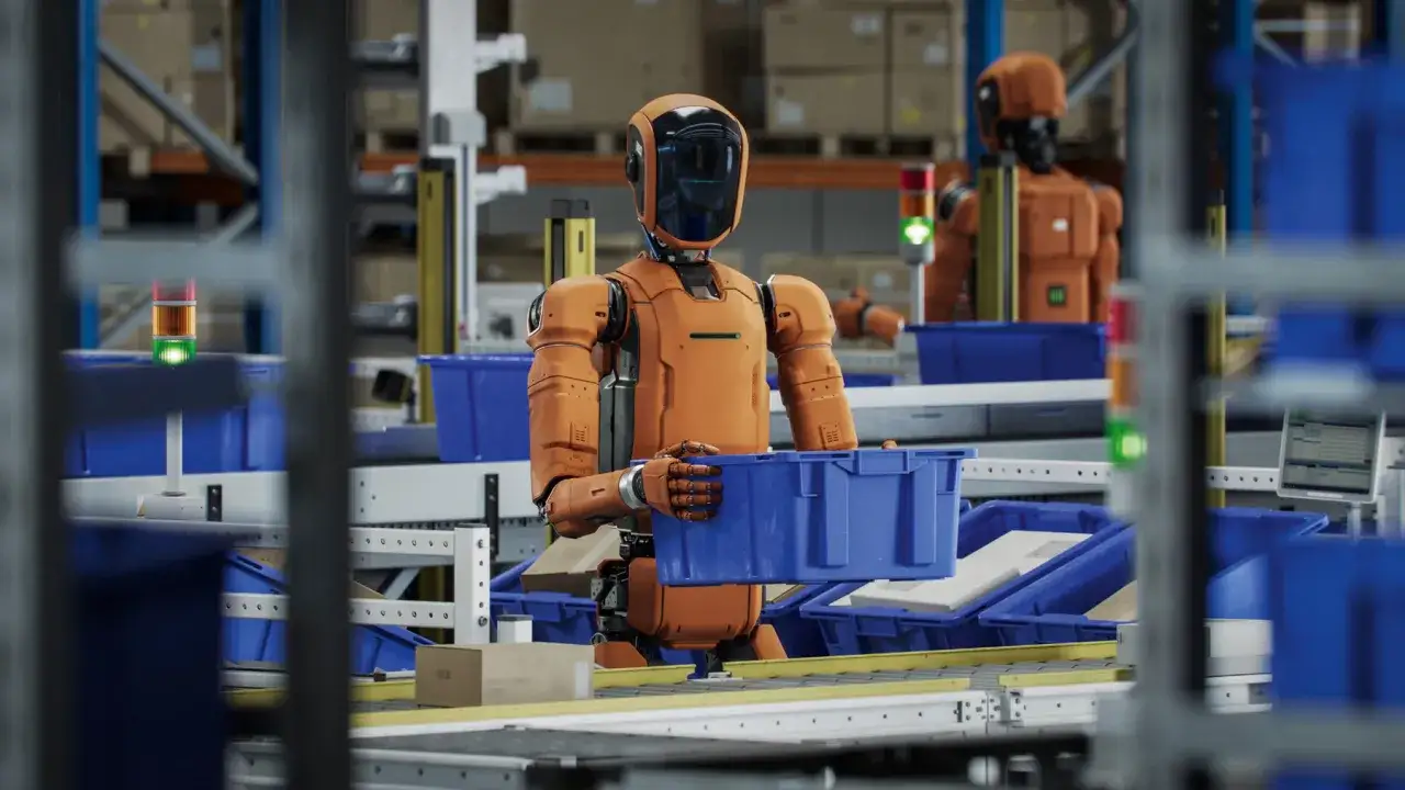 Pomarańczowy robot przenosi niebieskie pudełko w magazynie. To przykłady innowacji w logistyce, które usprawniają pracę.