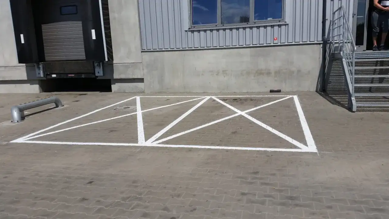 Parking z kopertą, czyli wyznaczone miejsce dla osób niepełnosprawnych, jest widoczne na brukowanym placu przed budynkiem magazynowym.