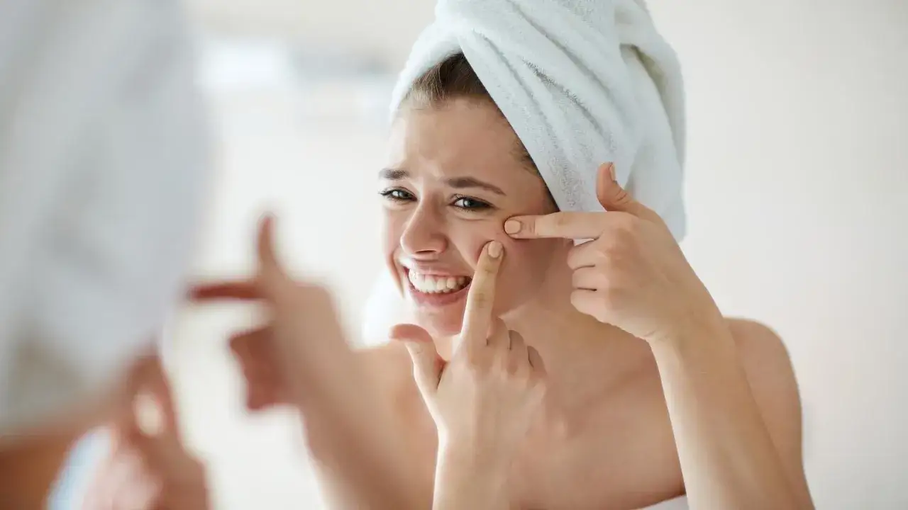 Kobieta z ręcznikiem na głowie, z grymasem na twarzy, pr&oacute;buje wyciskanie pryszczy przed lustrem.