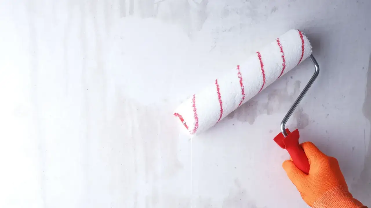 Ręka w pomarańczowej rękawicy nakłada wałkiem grunt pod tapetę flizelinową na szarą ścianę.