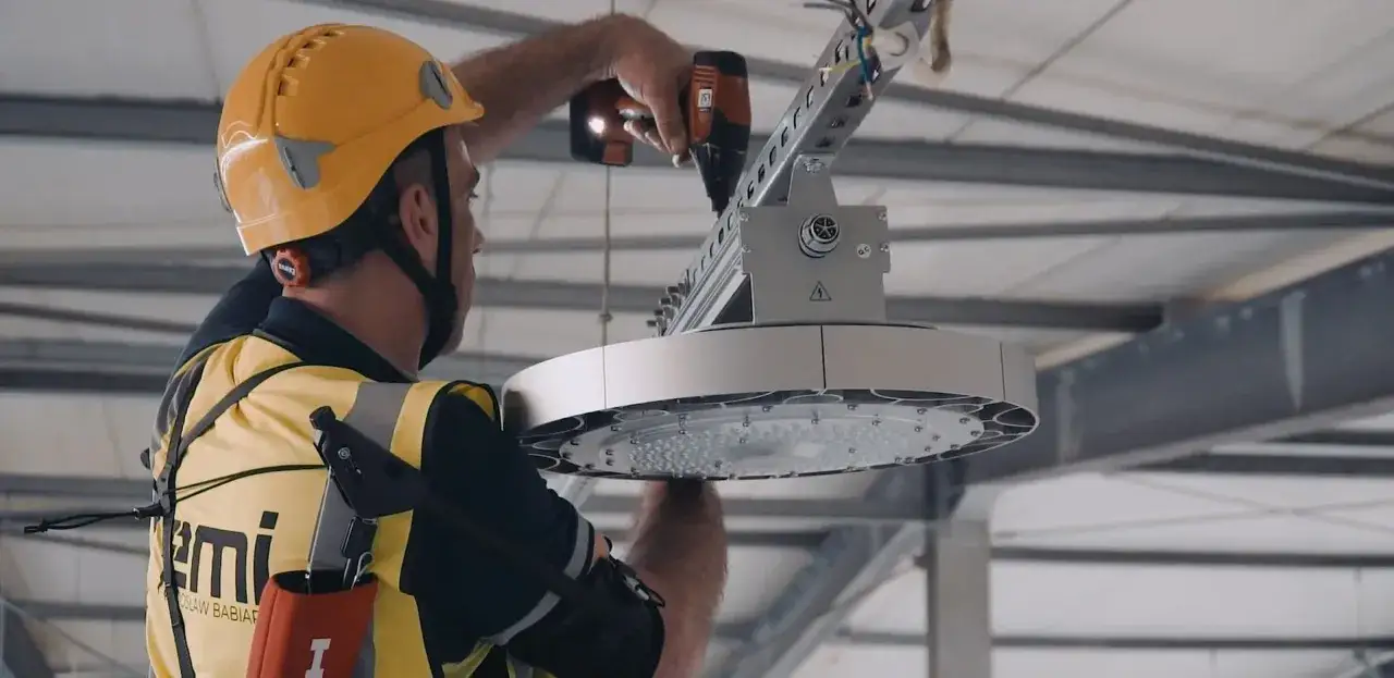 Elektryk w kasku montuje nowoczesne oświetlenie LED. To wymiana oświetlenia na led, remont czy modernizacja hali produkcyjnej.