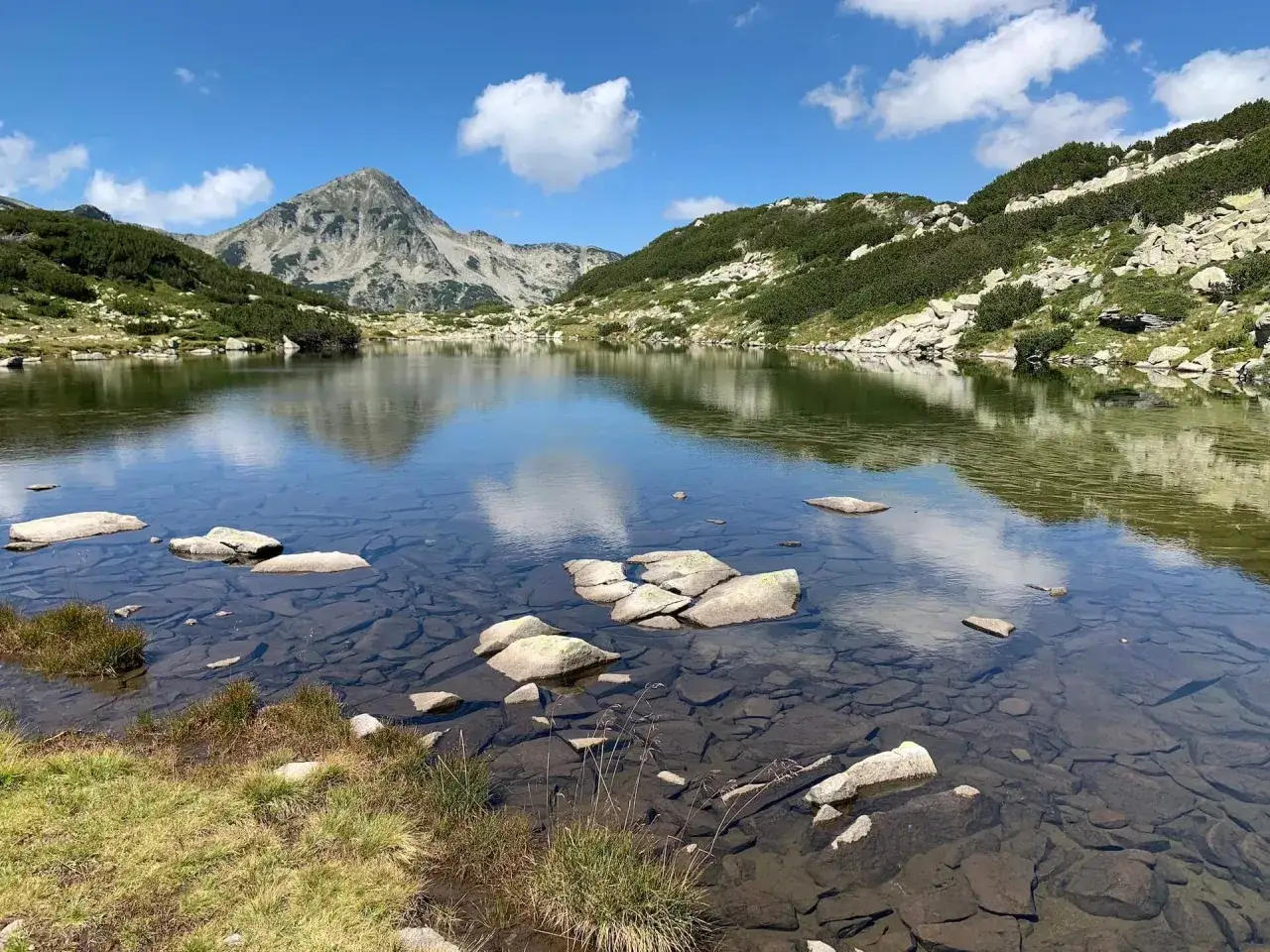 Spokojne jezioro odbija szczyty g&oacute;r Pirin. Kamienie na dnie są widoczne przez krystalicznie czystą wodę.