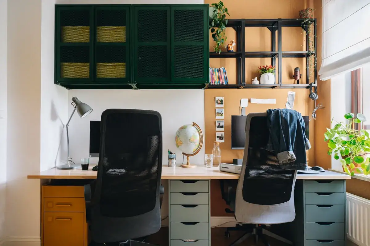 Dwa stanowiska pracy w domowym gabinecie. Biurko z dwoma krzesłami, monitorami, globusem i zielonymi szafkami nad nim.