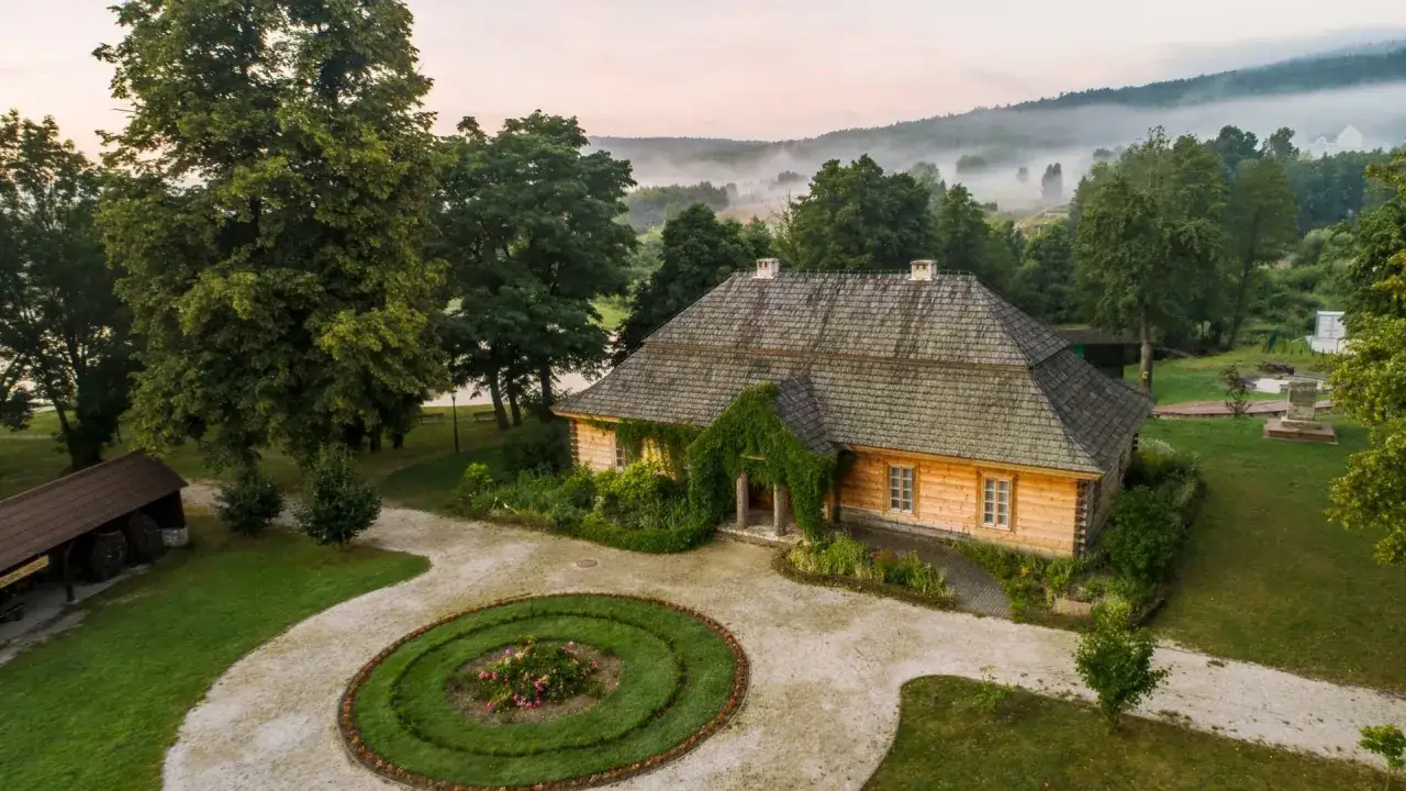 Drewniany dom otoczony zielenią, z mgłą w tle. Utopijny i realny obraz rzeczywistości, gdzie czas zatrzymał się w miejscu.