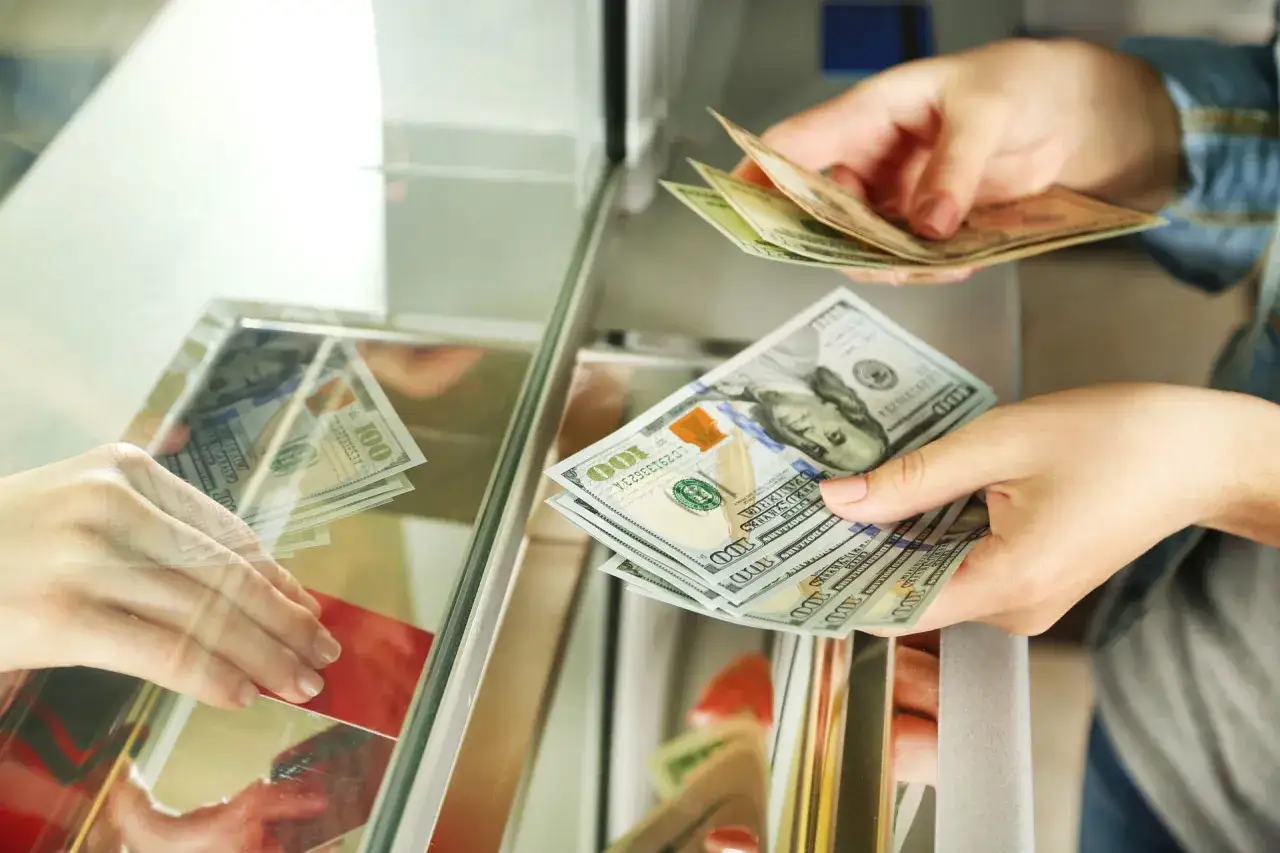 Ręce przekazują sobie banknoty dolarowe przez okienko. Tak, w banku można wymienić walutę.