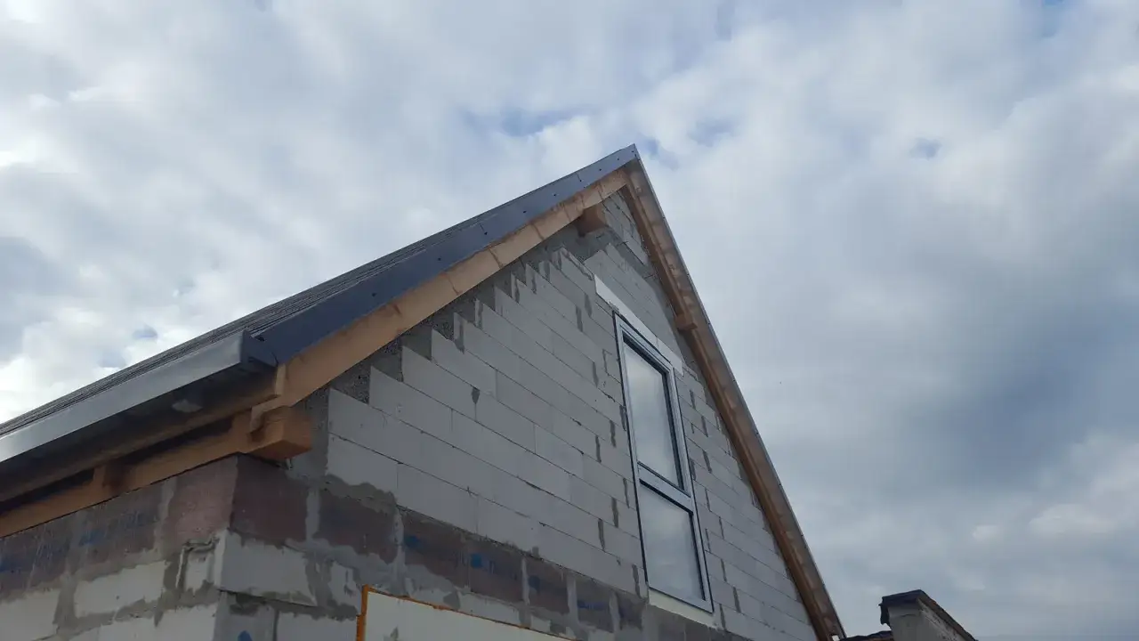 Rozpoczyna się wykończenie ściany szczytowej domu z bloczków. Widać fragment dachu i okno.