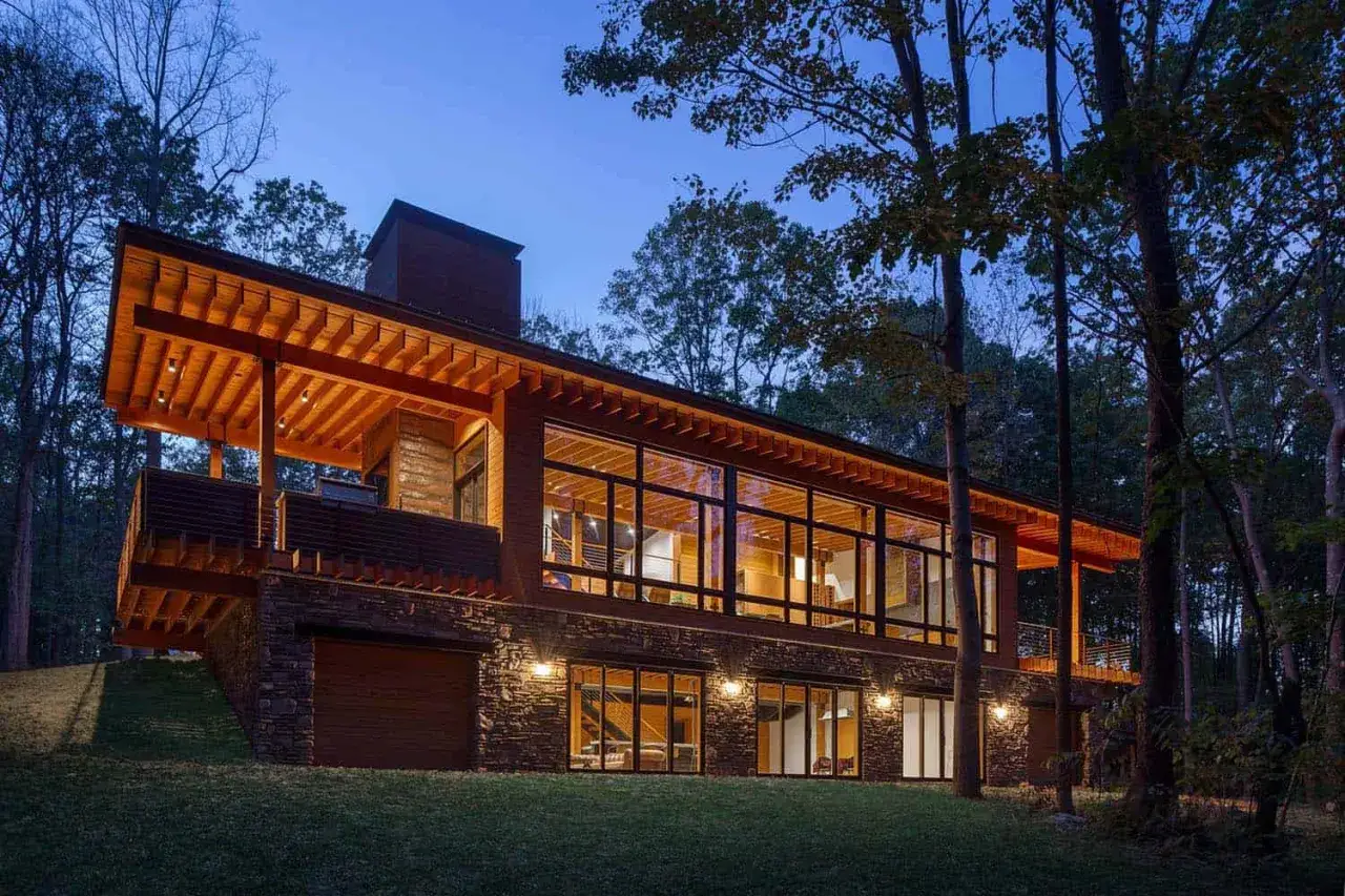 Nowoczesny dom drewniany w lesie, z dużymi oknami i tarasem, oświetlony wieczorem.