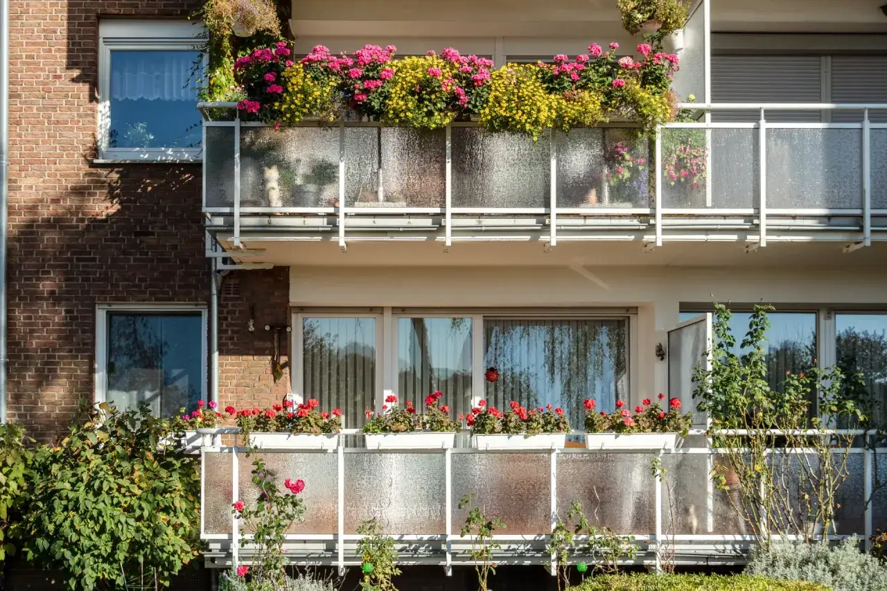 Kwiaty na balkonach w bloku, z różowymi i żółtymi pelargoniami na górze, a na dole czerwone róże.