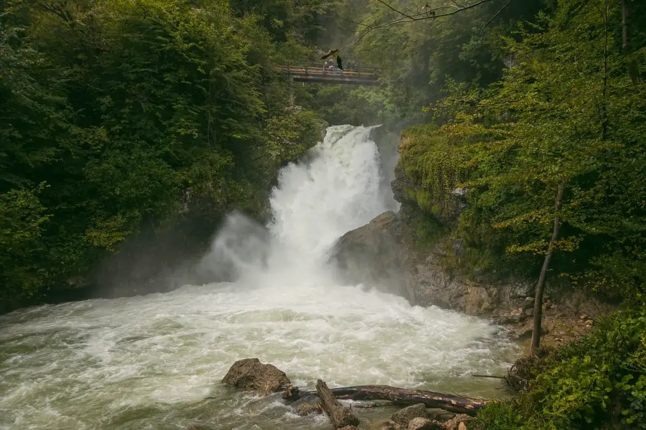 Potężny wodospad w wąwozie Vintgar, z mostkiem nad nim, gdzie widać ludzi. Woda spływa kaskadami do rwącego potoku.