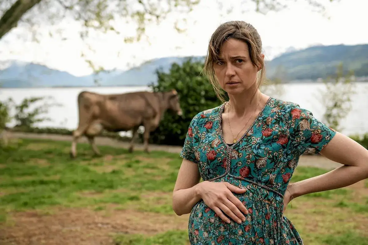 Agnieszka Grochowska w ciąży, z ręką na brzuchu, na tle łąki, jeziora i pasącej się krowy.