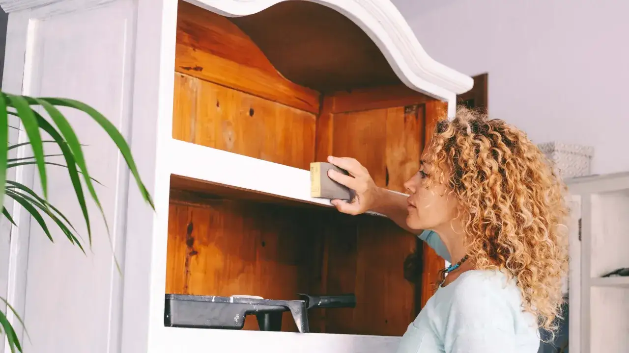 Kobieta szlifuje starą szafę, przygotowując ją do renowacji. Pokazuje, jak odnowić szafę z płyty, nadając jej nowe życie.