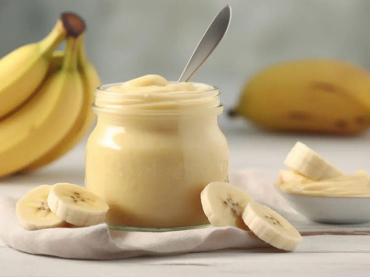 Gładkie puree bananowe w słoiku, idealne na biegunkę. Obok plasterki banana i kiść dojrzałych owoców.