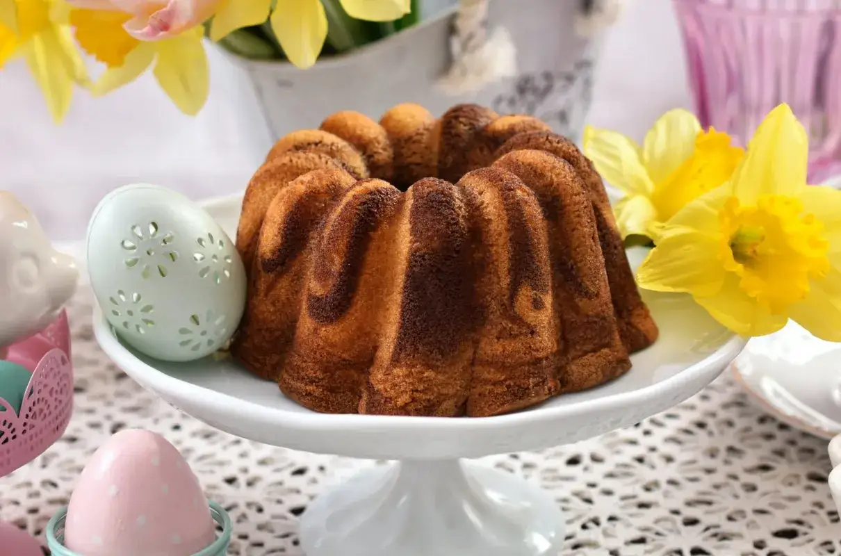 Wilgotna babka wielkanocna dwukolorowa, ozdobiona wzorem marmurkowym, prezentuje się apetycznie na białej paterze, otoczona wiosennymi kwiatami i pisankami.