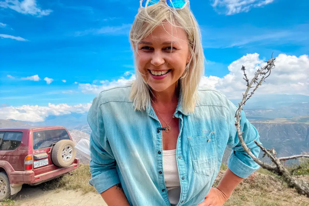 Agnieszka Kołodziejska, w wieku podróżniczki, uśmiecha się na tle gór i błękitnego nieba. Obok czerwony samochód terenowy.