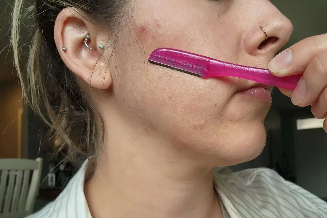 Kobieta wykonuje golenie twarzy u kobiet r&oacute;żową maszynką. Delikatne włoski znikają, pozostawiając gładką sk&oacute;rę.