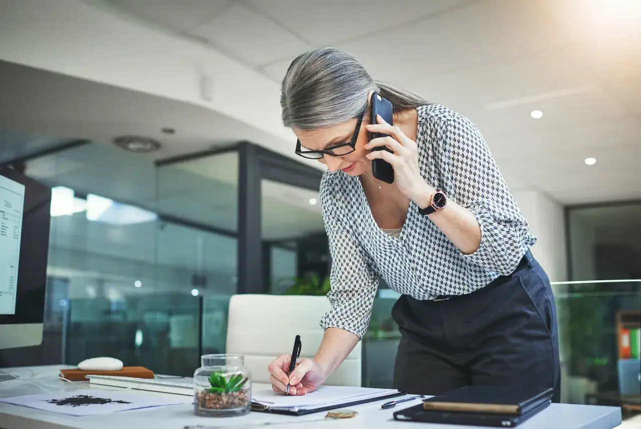 Kobieta w okularach rozmawia przez telefon i notuje. Dyskutuje o tym, ile czasu ma pracodawca na zgłoszenie pracownika do ZUS.