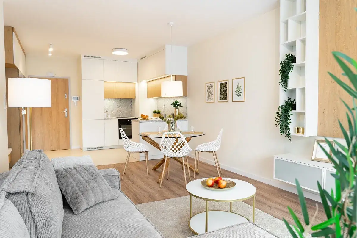 Nowoczesny salon z aneksem kuchennym, gdzie pakiety wykończenia pod klucz tworzą spójną całość. Szara sofa, okrągły stół z białymi krzesłami i funkcjonalna zabudowa kuchenna.