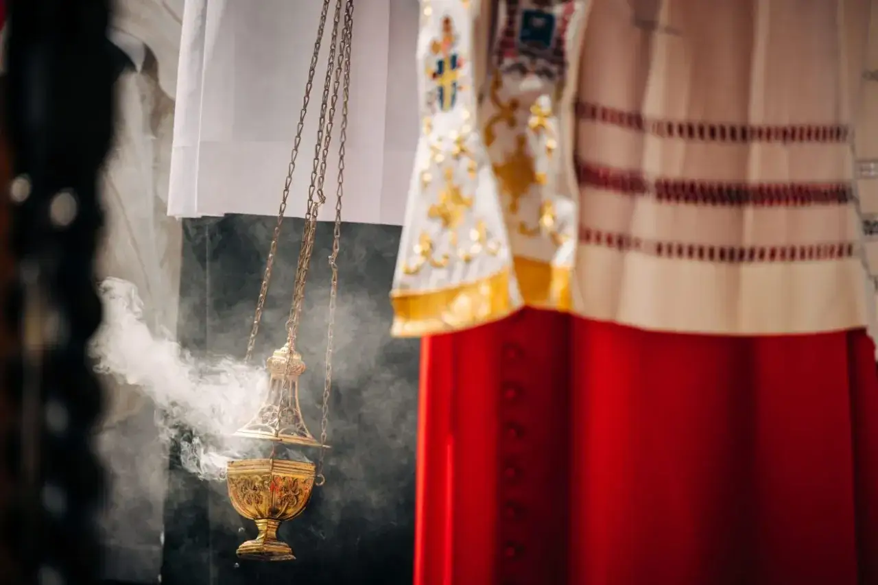 Dym z kadzielnicy unosi się podczas mszy wieczornej przed świętem. Złota kadzielnica wisi na łańcuszkach, a w tle widać szaty liturgiczne.