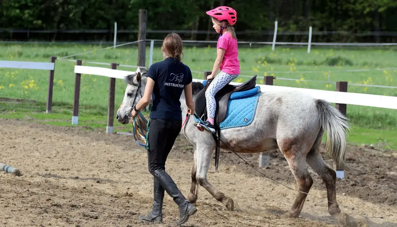 Instruktorka prowadzi młodą adeptkę jazdy konnej na siwym koniu na terenie klubu jeździeckiego Profit.